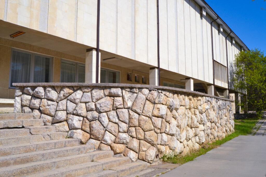 Modernes Gebäude und Stützmauer aus Naturstein, mit Mauerabdeckplatten darauf und eine Treppe mit Stufen.