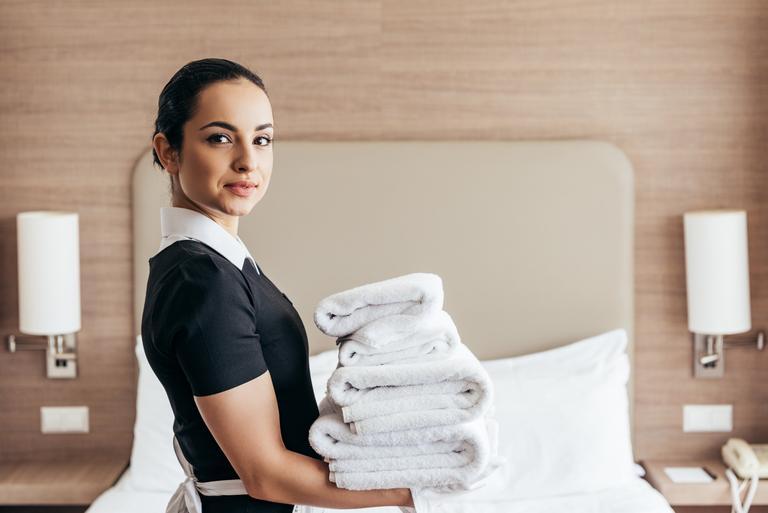 Lächelndes Zimmermädchen, das einen Stapel gefalteter Handtücher in der Nähe des Bettes hält und in die Kamera schaut, in einem Hotelzimmer